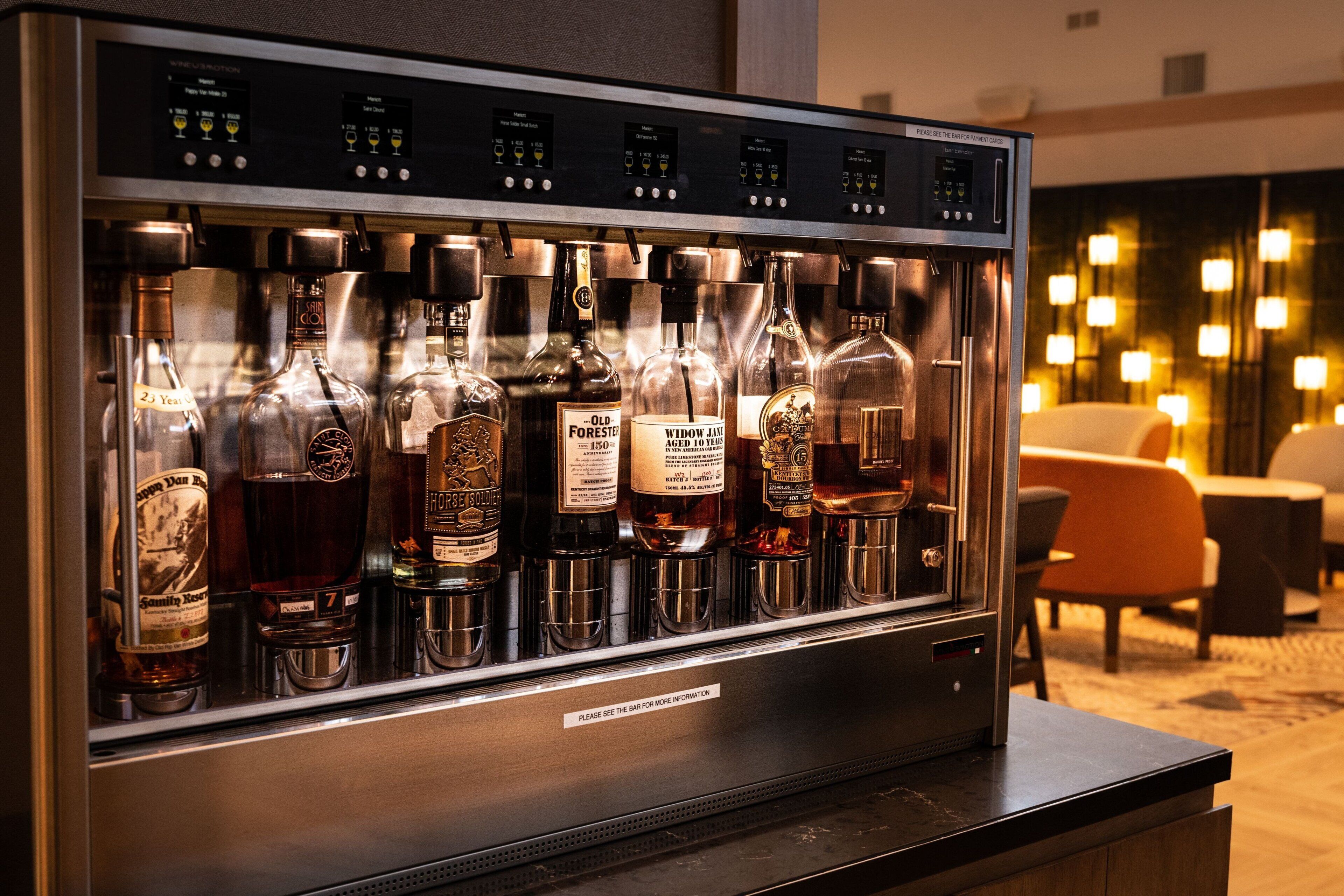 Een drankkoelkast gevuld met diverse flessen drank, een digitaal bedieningspaneel en een bordje met de tekst: voor meer informatie, neem contact op met de bar.