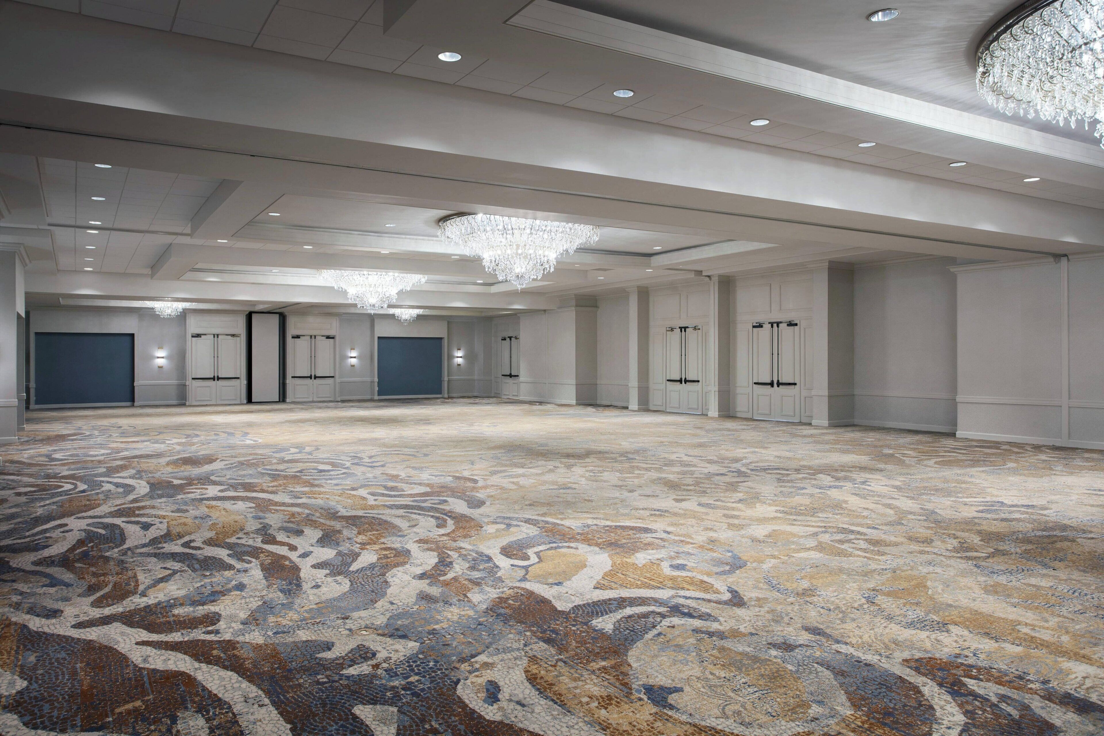 A spacious, well-lit room with a patterned carpet, multiple doors, and a large chandelier.