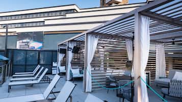 A rooftop lounge area with sun loungers, a covered seating area, and a view of the cityscape.