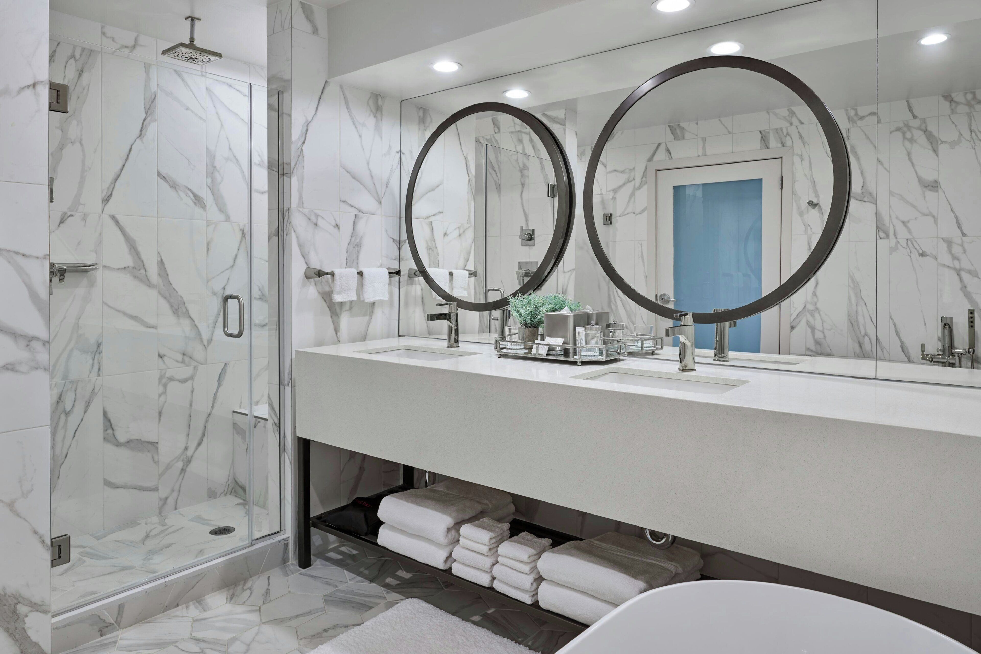 Un baño moderno con ducha envuelta en vidrio, un espejo grande con dos marcos circulares y un tocador blanco con lavamanos y espacio para toallas.
