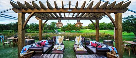Ein Außenbereich mit Holztischen und Ledersofas unter einer Pergola, mit Blick auf einen Golfplatz.