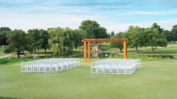 Campo de golf con sillas blancas dispuestas en filas, una pérgola de madera y un estanque al fondo.