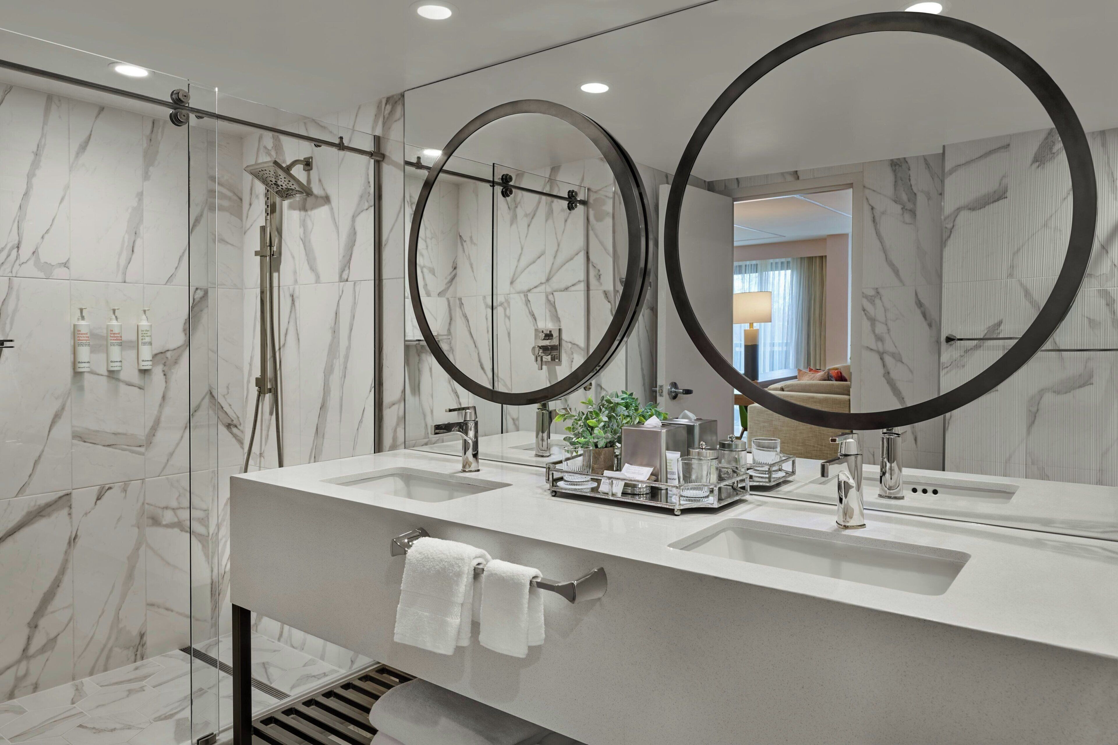 A modern bathroom with two sinks, a shower, and a large mirror with a circular frame.