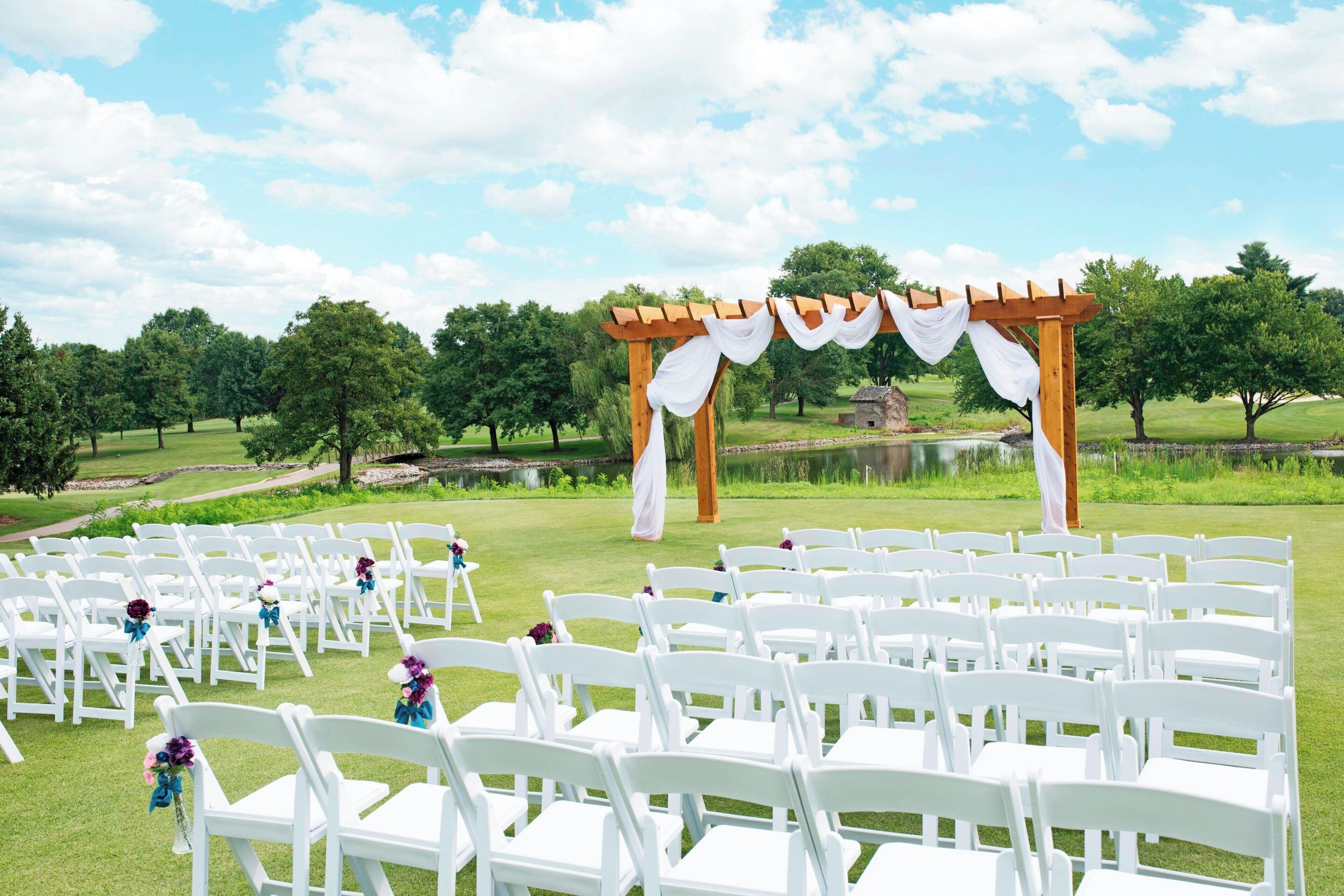 A wedding ceremony setup with white chairs arranged in rows, a wooden arch with white drapery, and a pond in the background.