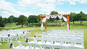 Preparación para una ceremonia de boda con sillas blancas dispuestas en filas, un arco de madera con cortinas blancas y un estanque al fondo.