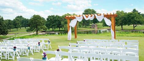 A wedding ceremony setup with white chairs arranged in rows, a wooden arch with white drapery, and a pond in the background.