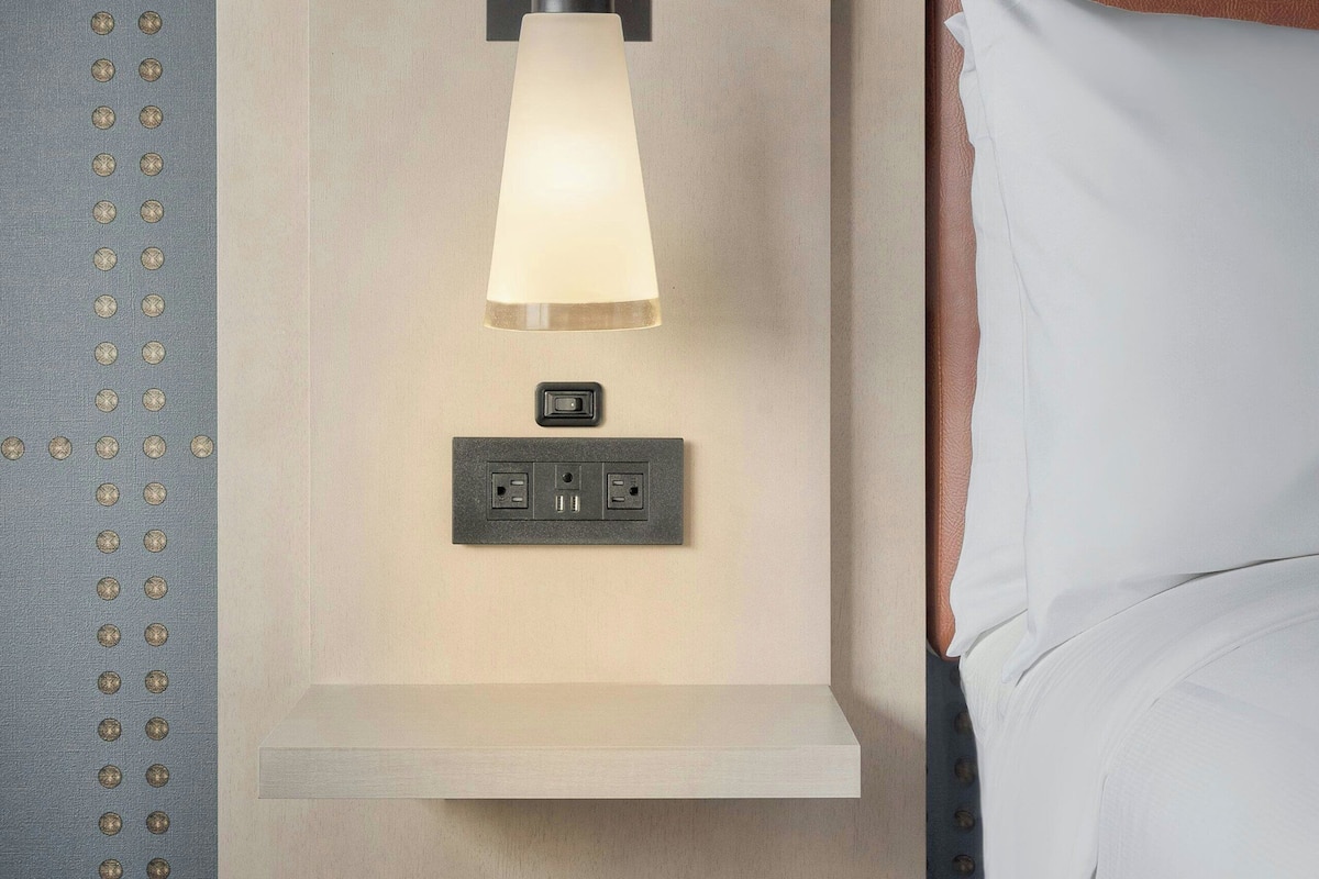 A modern hotel room with a bed, a wall-mounted light fixture, and a power outlet.