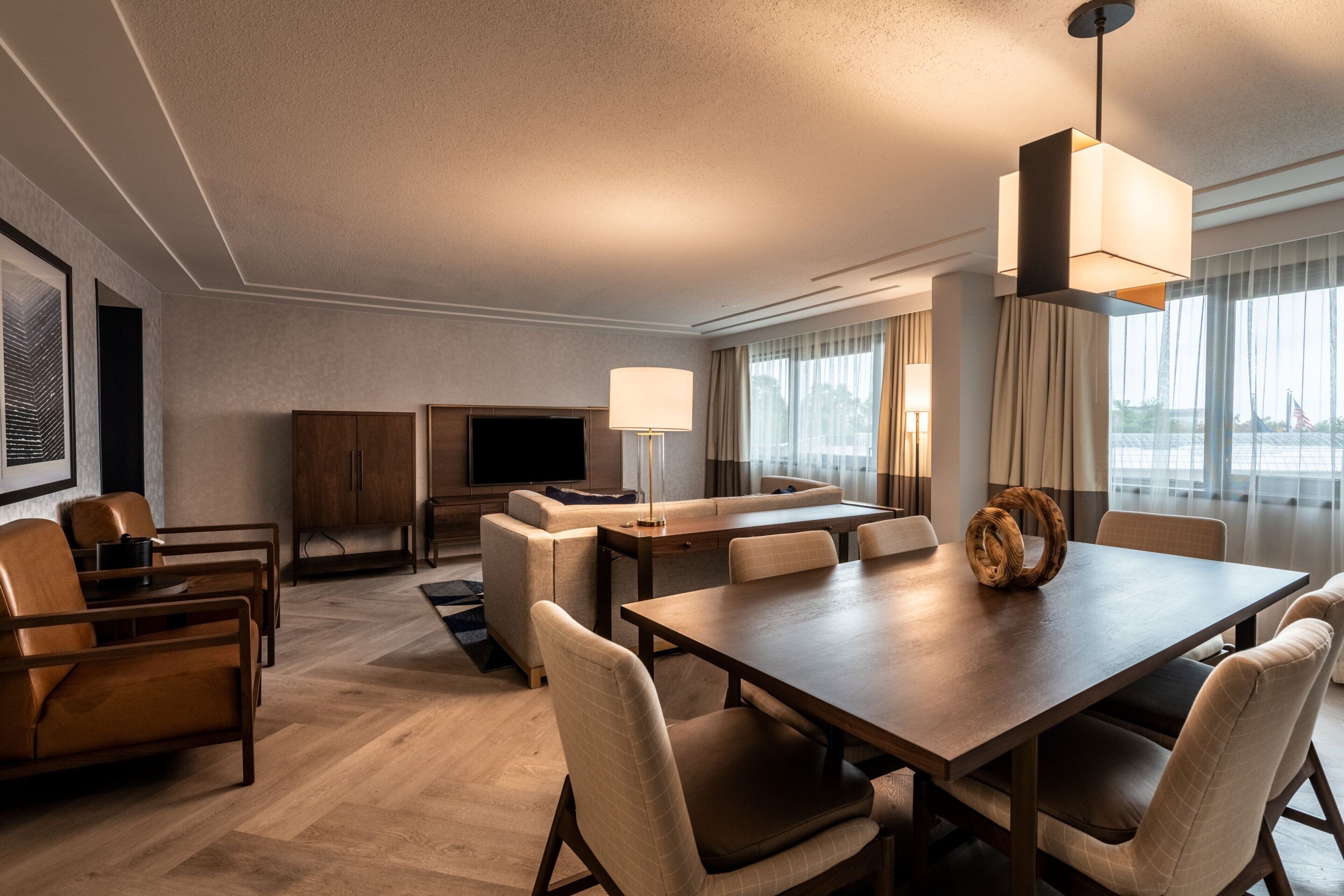 Habitación de hotel con comedor, sofá, televisor y un amplio ventanal con cortinas.