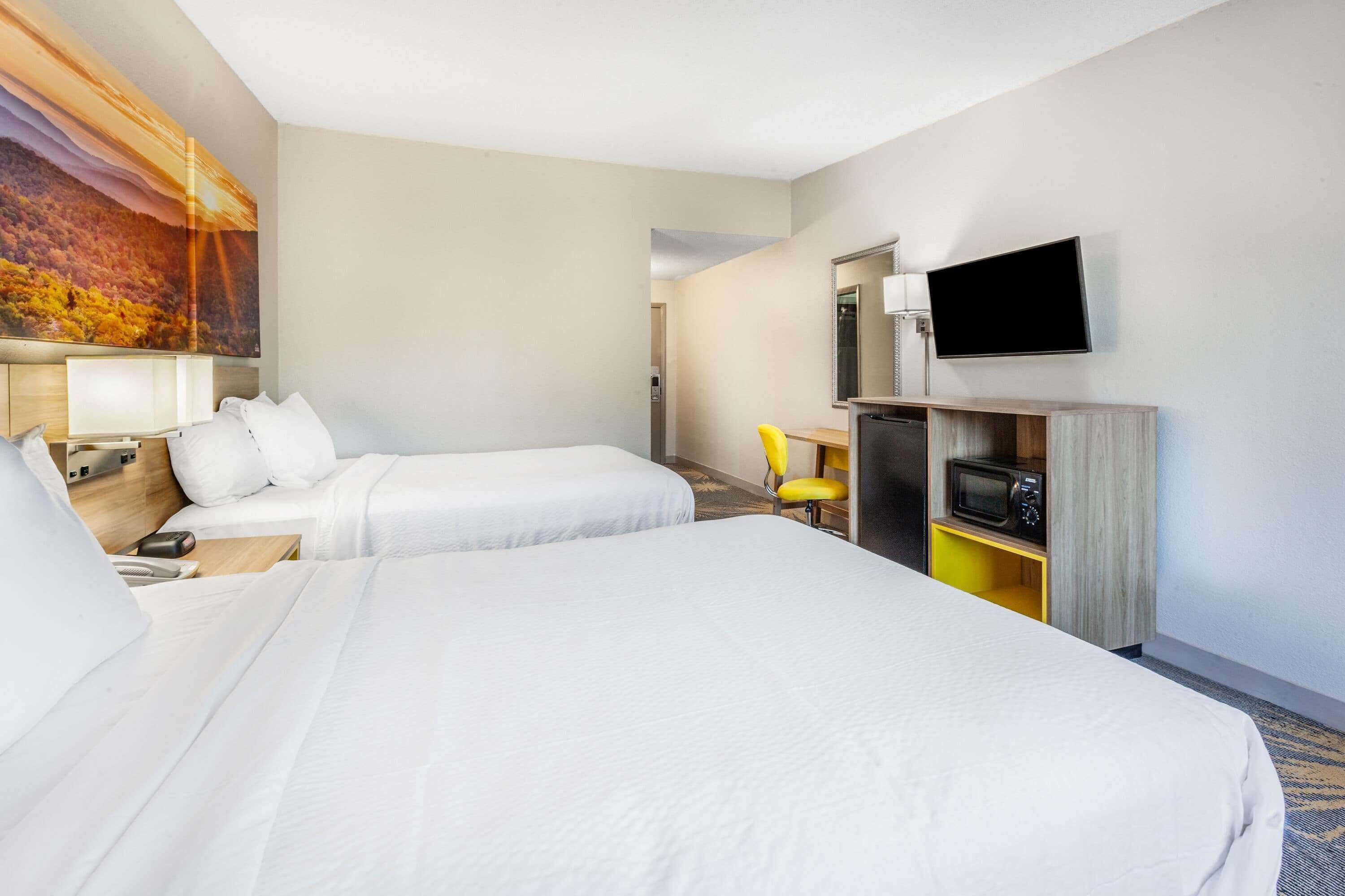 A hotel room with two beds, a desk with a chair, a flat-screen TV, a microwave, and a painting on the wall.