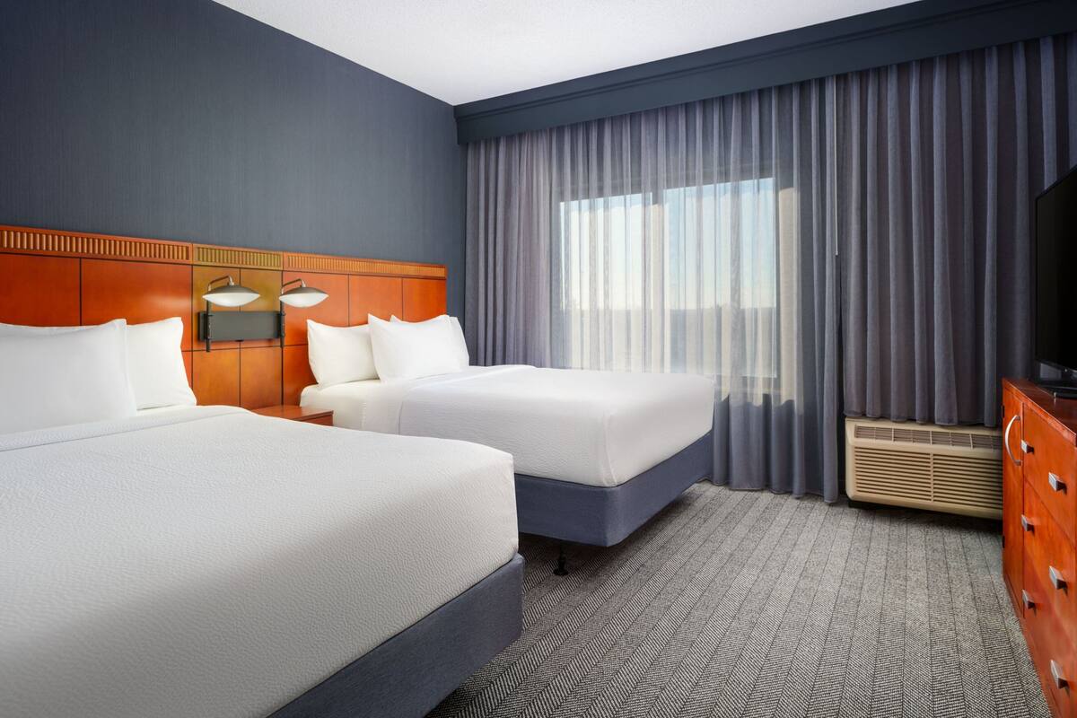 A hotel room with two beds, a wooden headboard, a large window with curtains, a flat-screen TV, and a dresser.