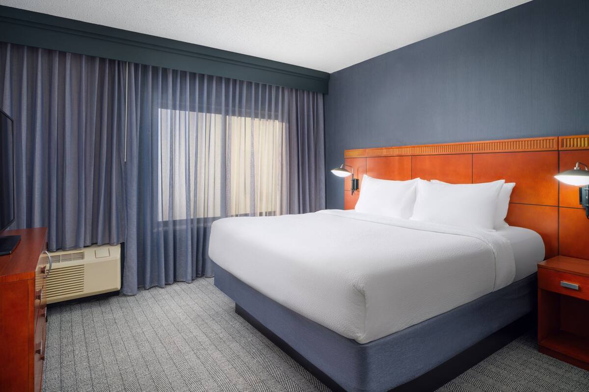 A hotel room with a large bed, a wooden headboard, two bedside lamps, a wooden nightstand, and a large window with curtains.