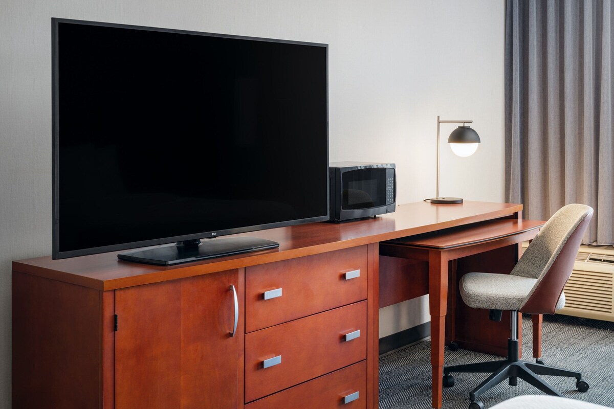 A hotel room with a wooden desk, a flat-screen TV, a microwave, a desk lamp, and a cushioned chair.