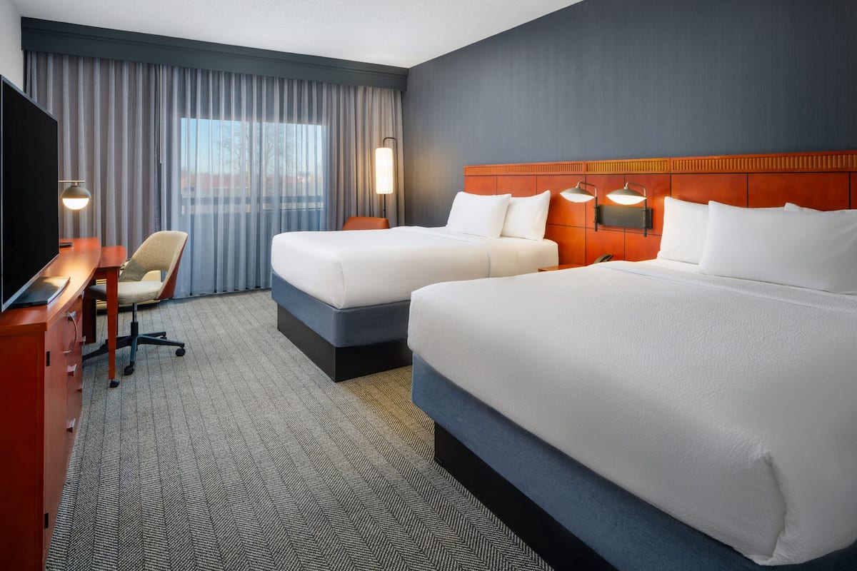 A hotel room with two beds, a desk, a chair, and a TV.
