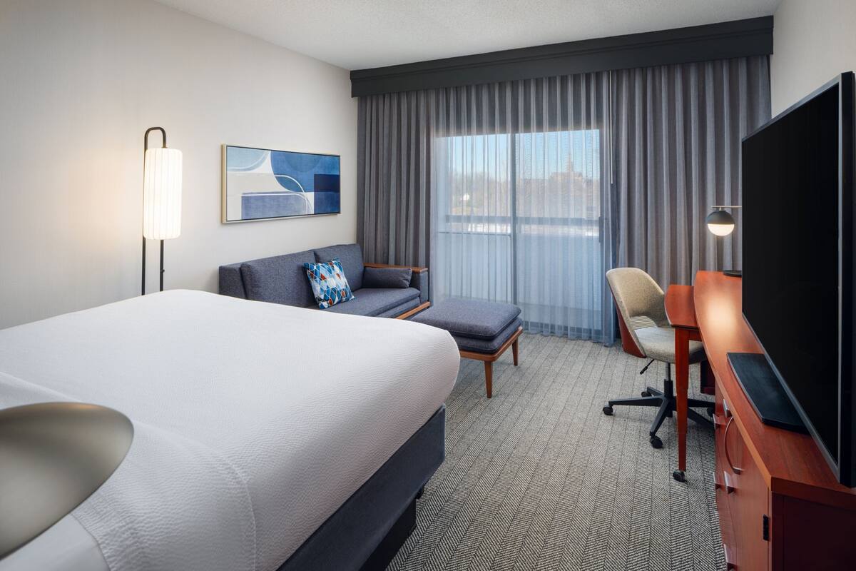 A hotel room with a large bed, a sofa, a desk with a TV, a chair, and a window with curtains.
