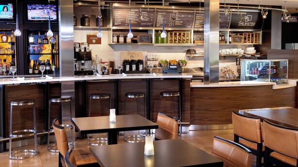 A modern bar area with a counter, stools, and tables. There are menu boards above the counter and a television screen displaying a sports event. - Courtyard by Marriott Columbus Worthington (Columbus)