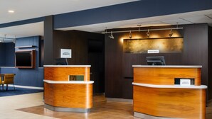 A hotel reception area with two wooden reception desks, a seating area with a table and chairs, and a dark wall with a decorative panel. - Courtyard by Marriott Columbus Worthington (Columbus)