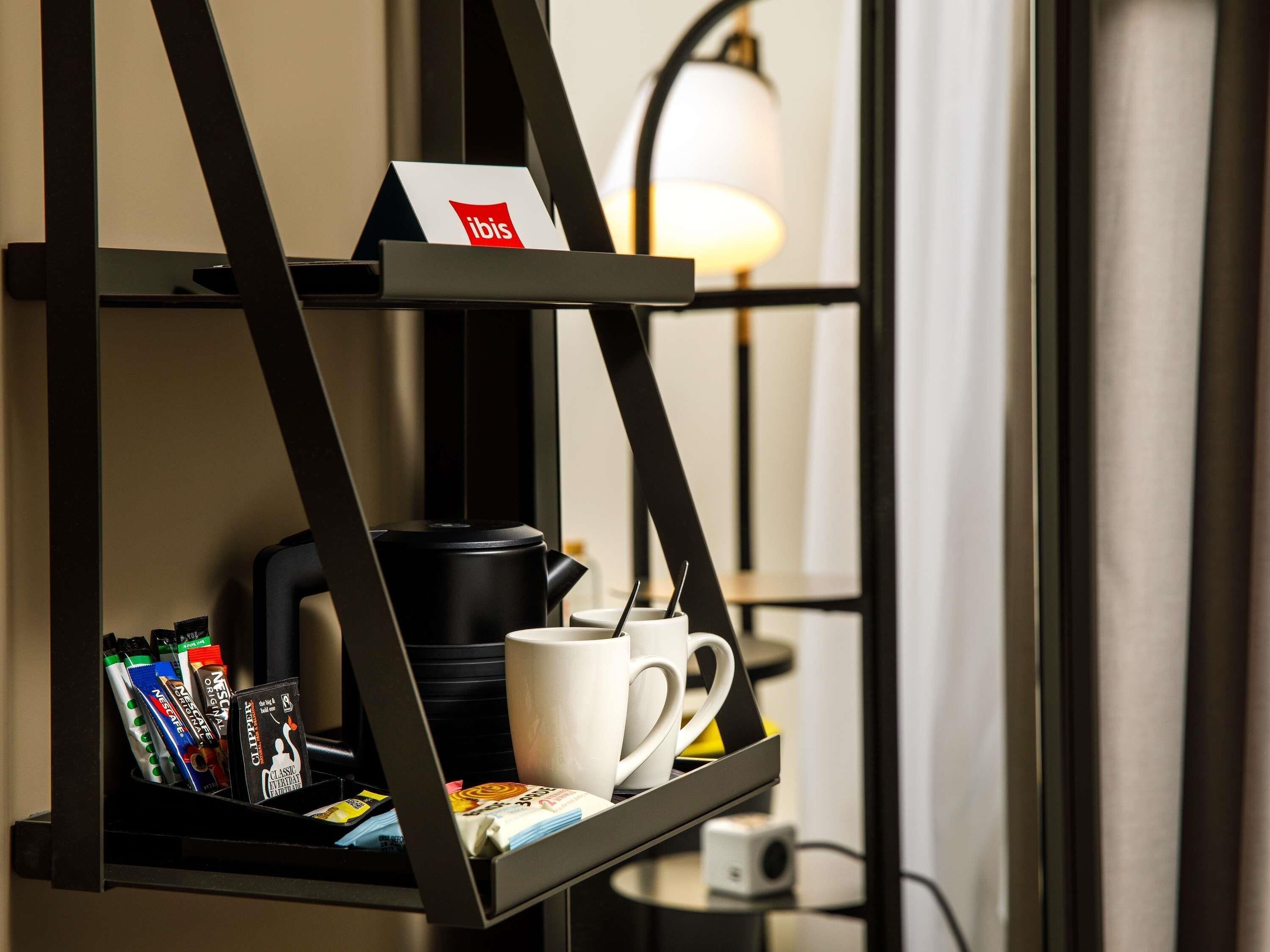 Una repisa negra con una cafetera, una taza y una tarjeta que dice "ibis" en un entorno hotelero.