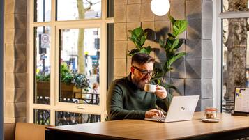 A person working on a laptop at a wooden table in a cafe with large windows and indoor plants.