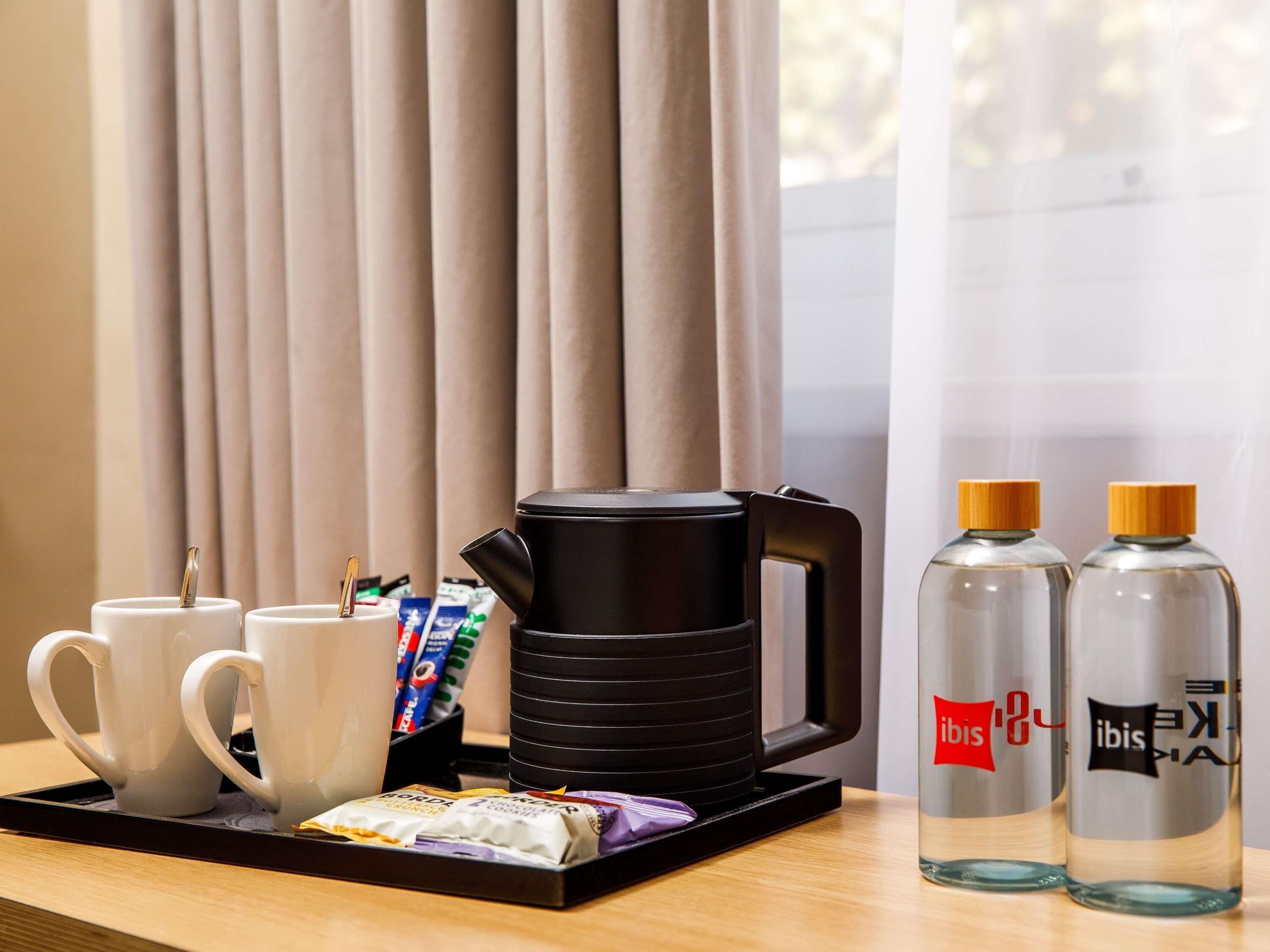 A tray with two white mugs, a black electric kettle, a stack of black plates, a packet of snacks, and two bottles labeled 'ibis.'