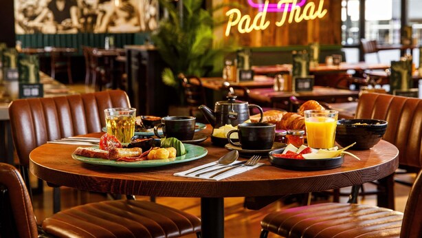 En restaurangmiljö med en neonreklamskylt som visar texten ”Pad Thai”, ett bord med frukosträtter och läderstolar.