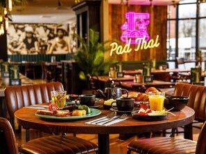 A restaurant setting with a neon sign reading 'Pad Thai,' a table with breakfast items, and leather chairs. - ibis London Greenwich (London)