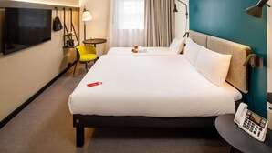 Ett hotellrum med en stor säng, en telefon vid sängen, en tv, ett litet bord, en stol och ett fönster med gardiner.