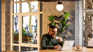 En person sitter vid ett träbord på ett kafé och arbetar på en bärbar dator. Kaféet har stora fönster och många krukväxter.
