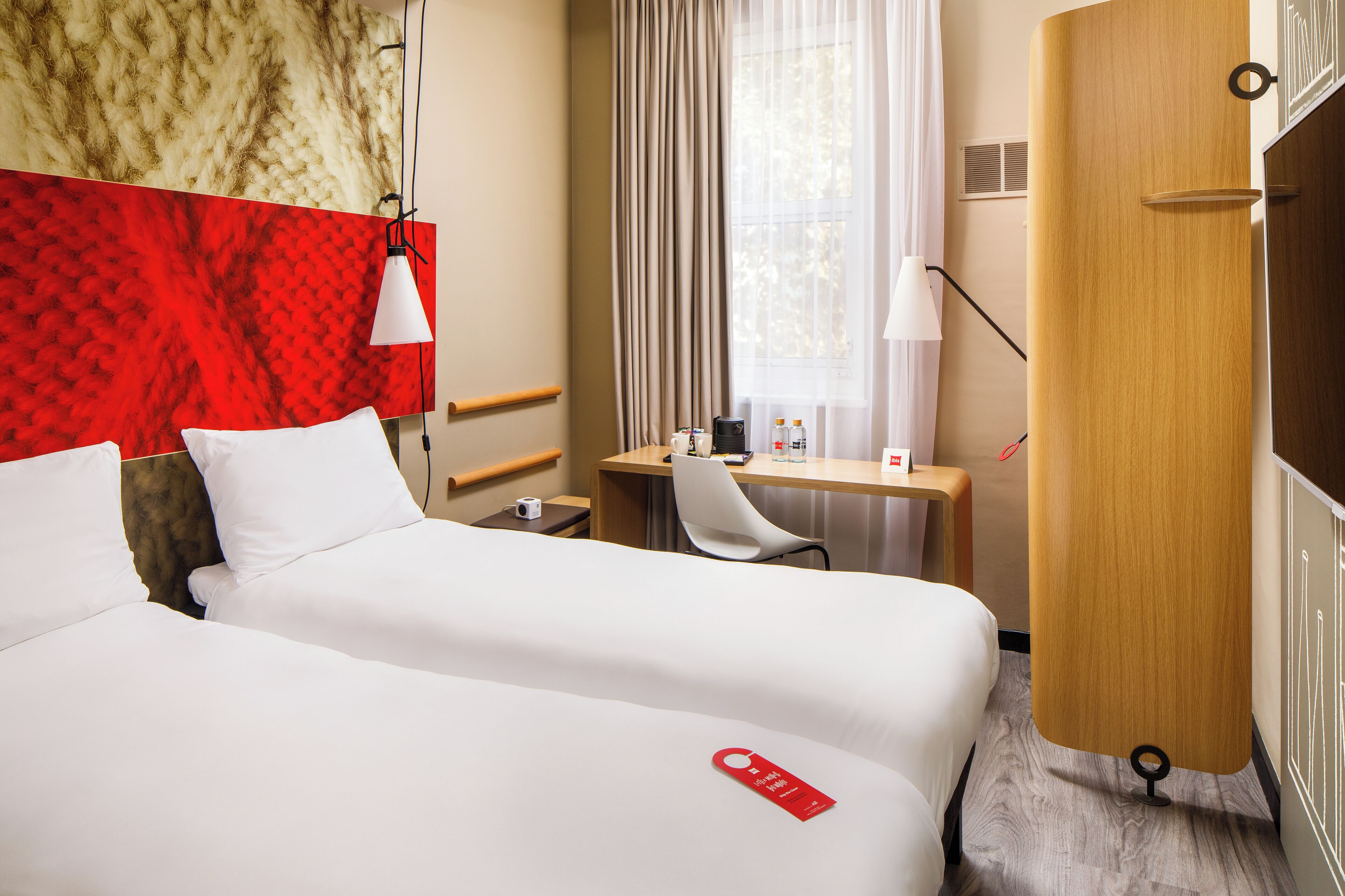 A hotel room with two beds, a desk, a chair, a window with curtains, and a wooden wardrobe.