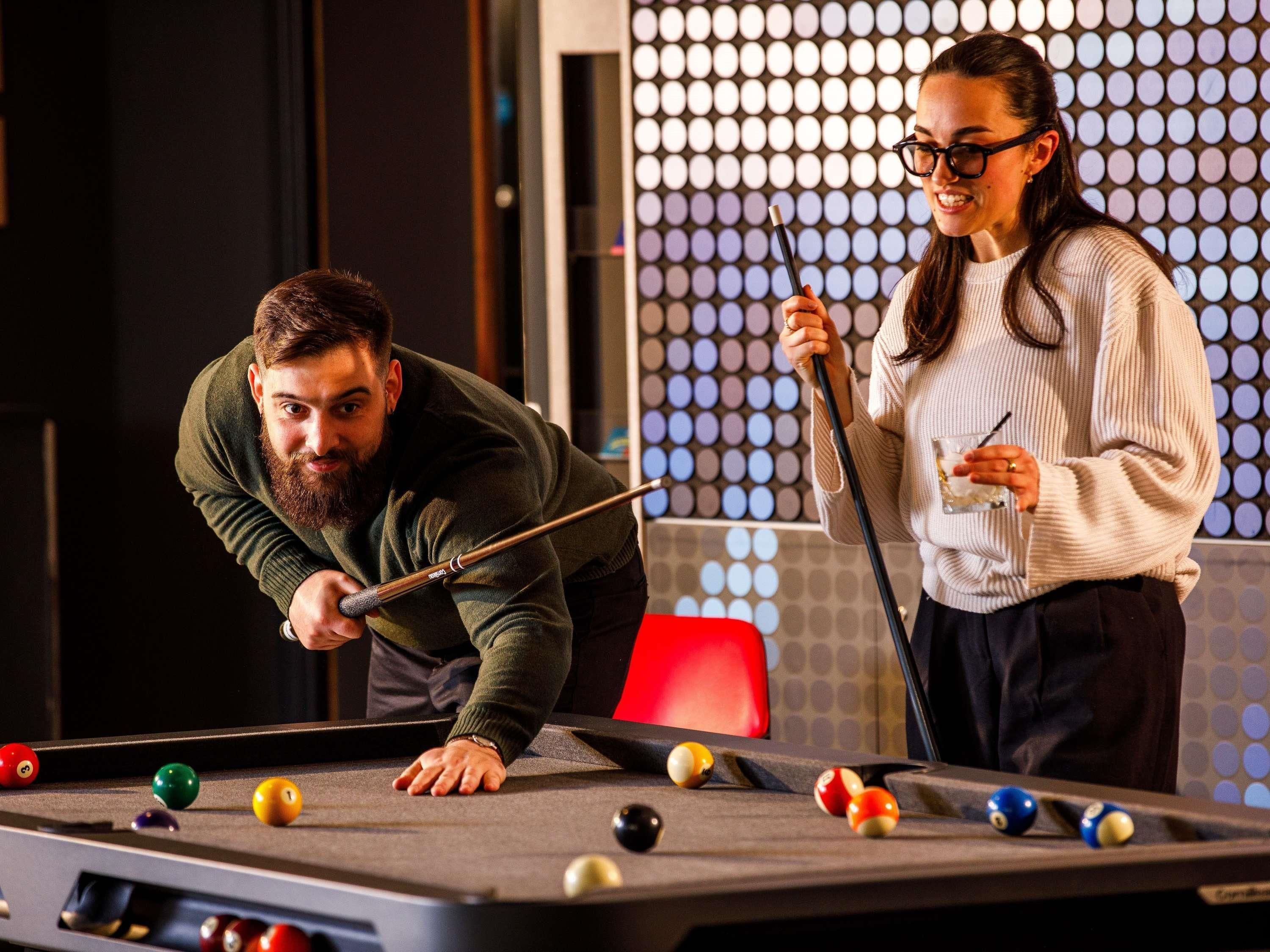 Une table de billard avec des billes et des queues, une chaise rouge et un mur à motifs en arrière-plan.