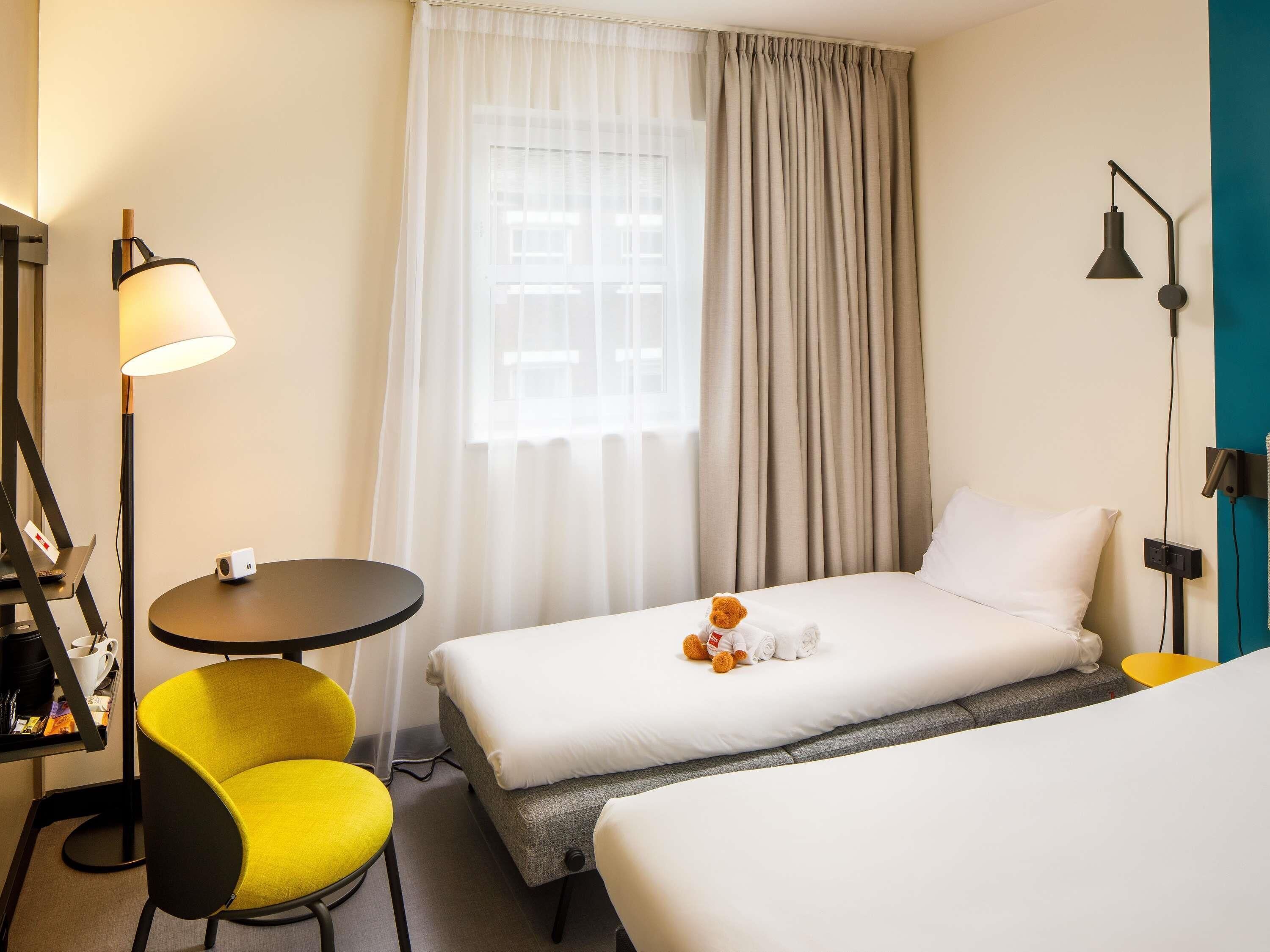 A hotel room with two beds, a desk with a lamp, a chair, and a window with curtains.