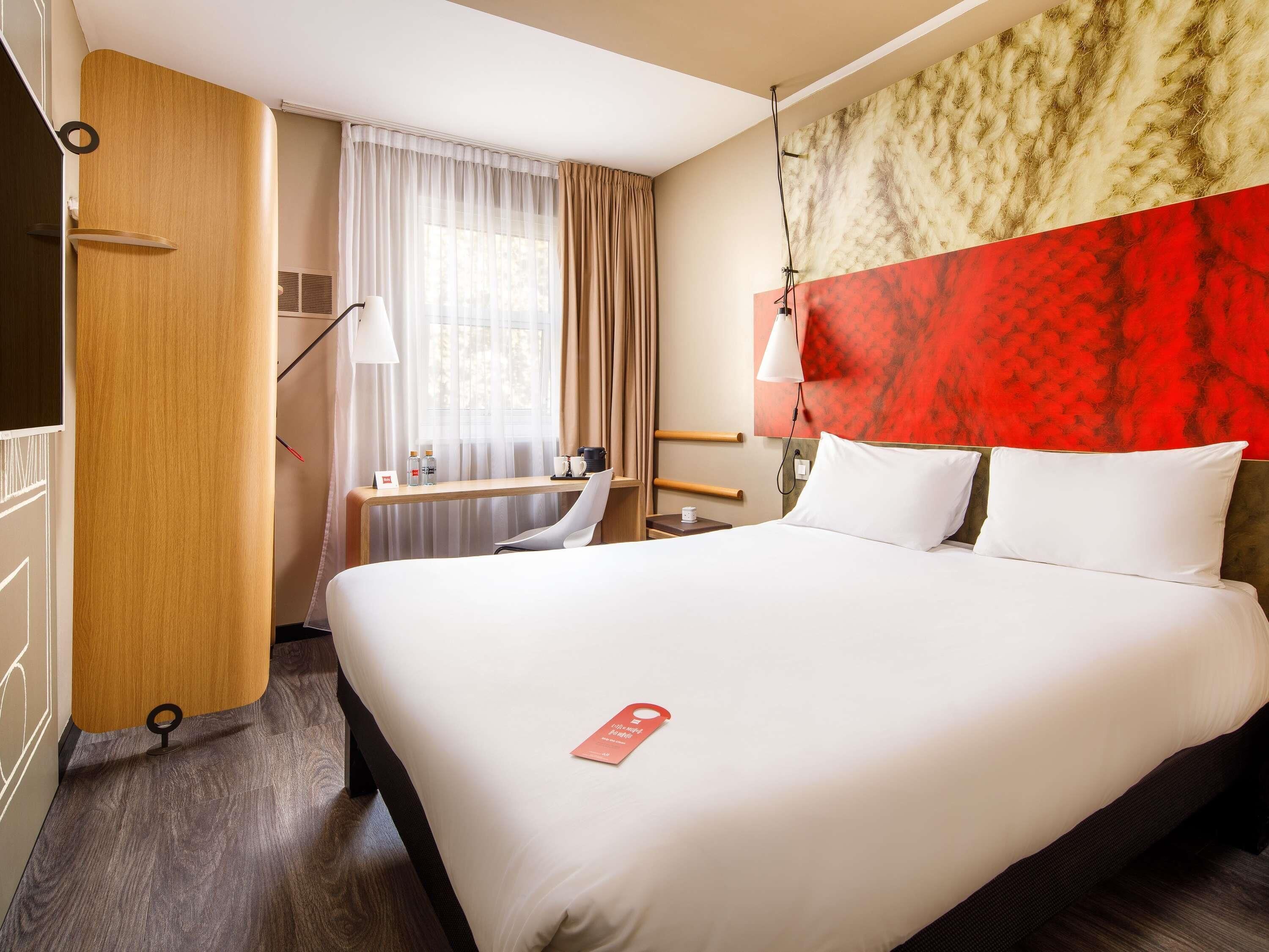 A hotel room with a bed, a desk, a chair, a lamp, and a window with curtains.