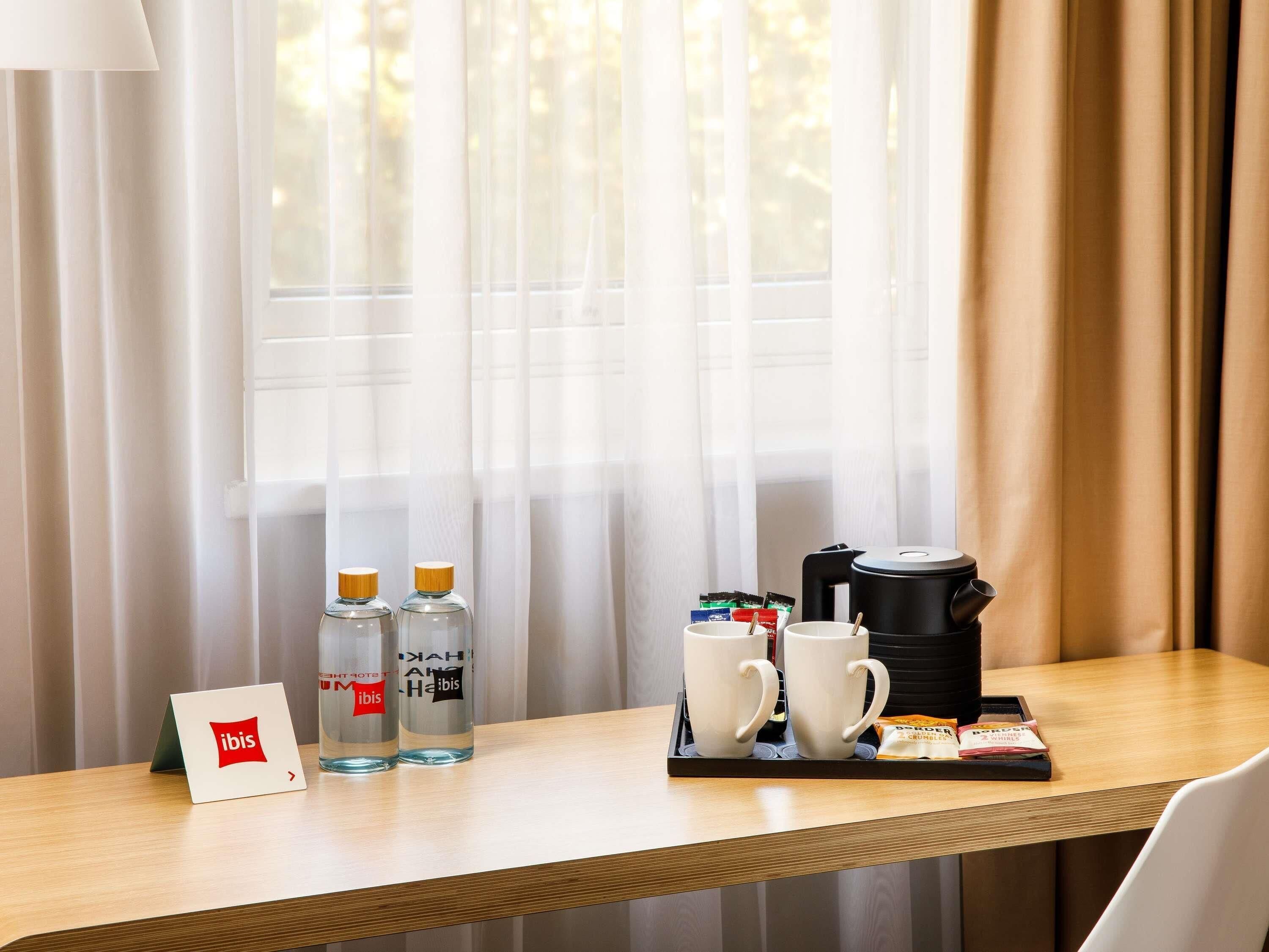Et hotelværelse med et træbord dækket med to vandflasker, to hvide krus, en kaffemaskine og en bakke med sukker og fløde.