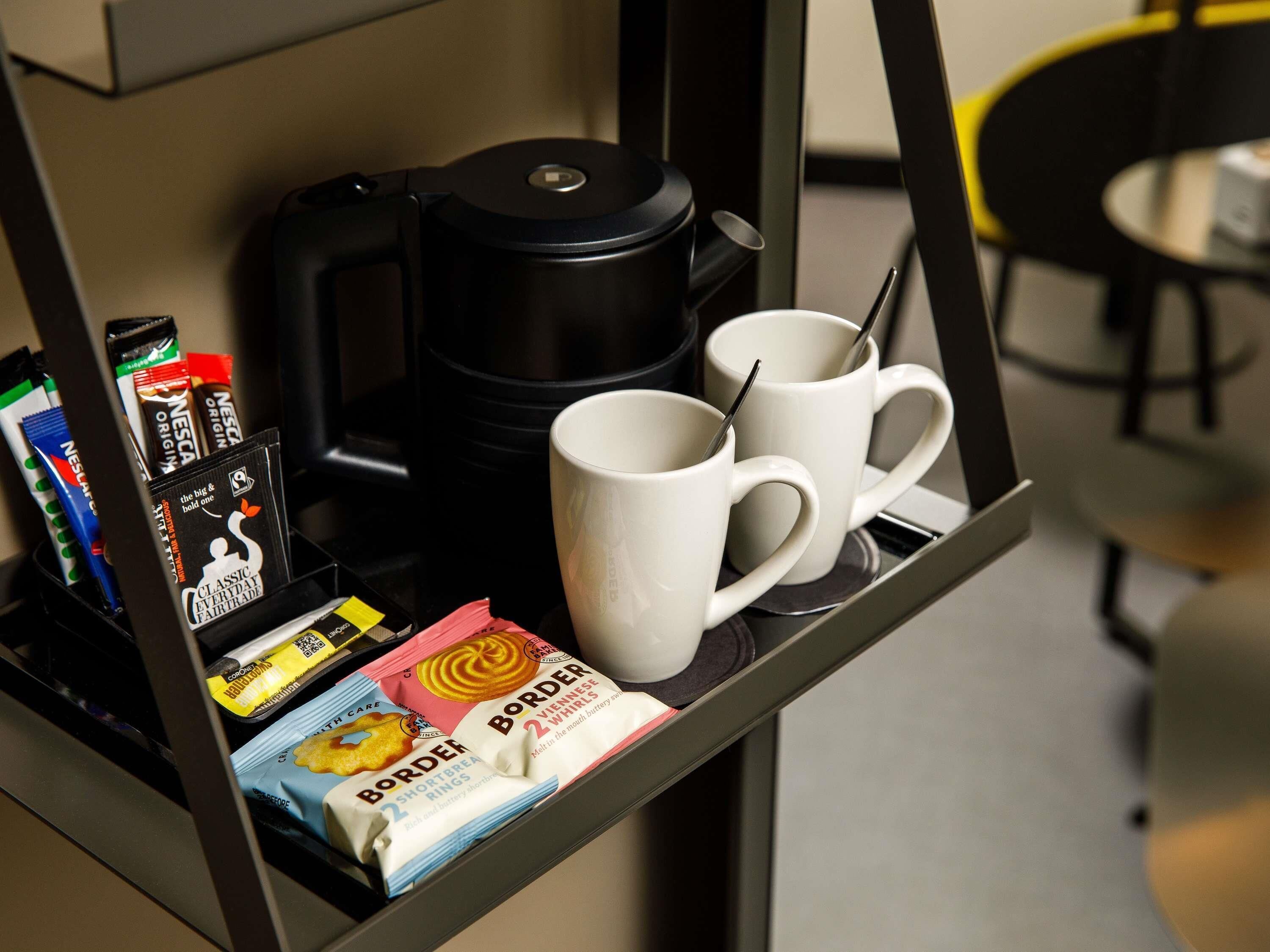 Un coin café équipé d’une machine à café, de deux tasses blanches et de divers en-cas, notamment des dosettes Nescafé et des barres de chocolat Border.