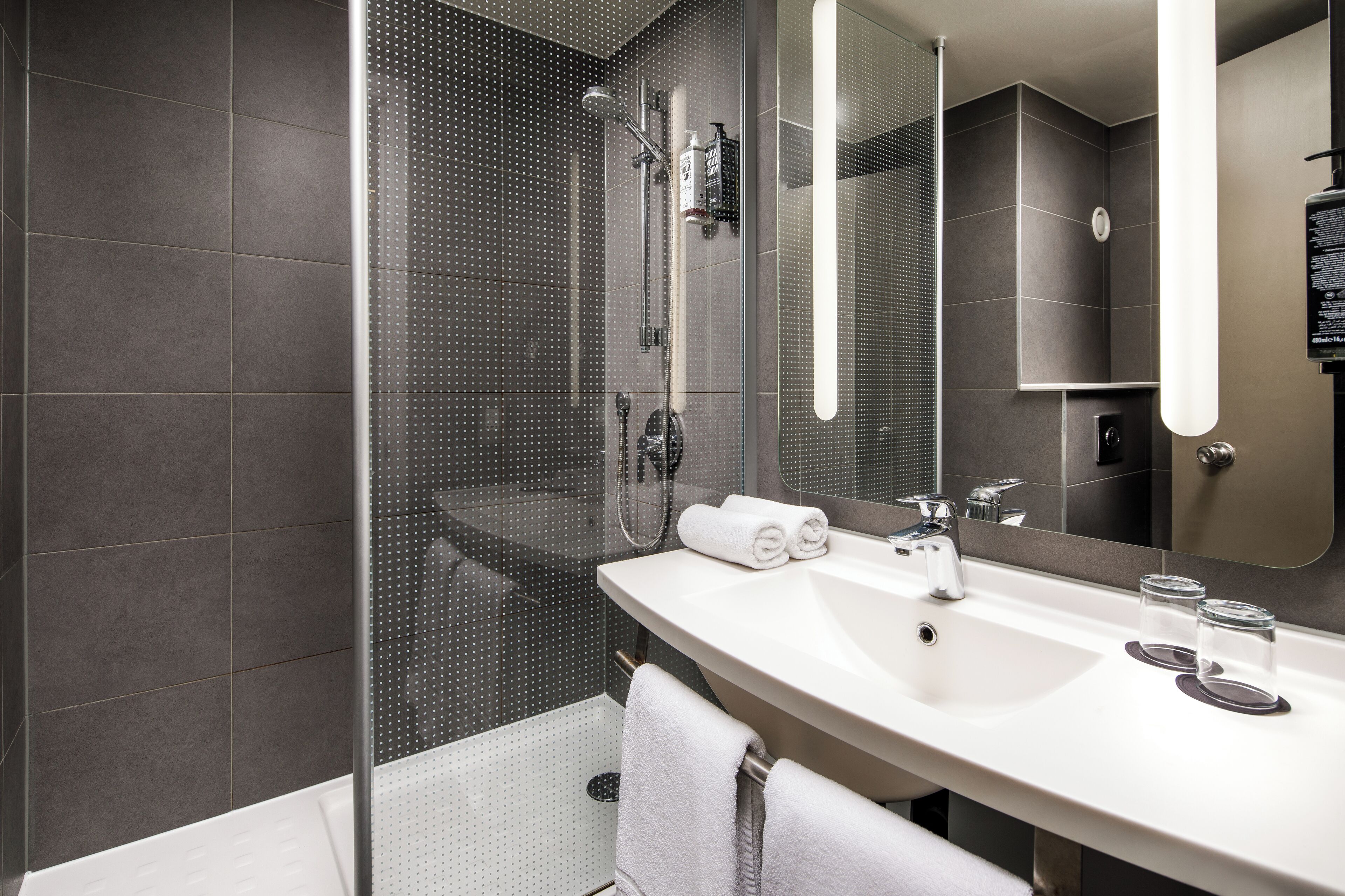 Ein modernes Badezimmer mit einer gläsernen Duschkabine, einem weißen Waschbecken mit zwei Spiegeln und einem Handtuchhalter.