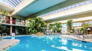An indoor swimming pool with a clear view of the sky through the ceiling, surrounded by lush greenery and a stone waterfall feature.