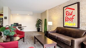 A hotel lobby with a Red Roof Plus sign, a sofa, a coffee table, a desk with a computer, and a plant.