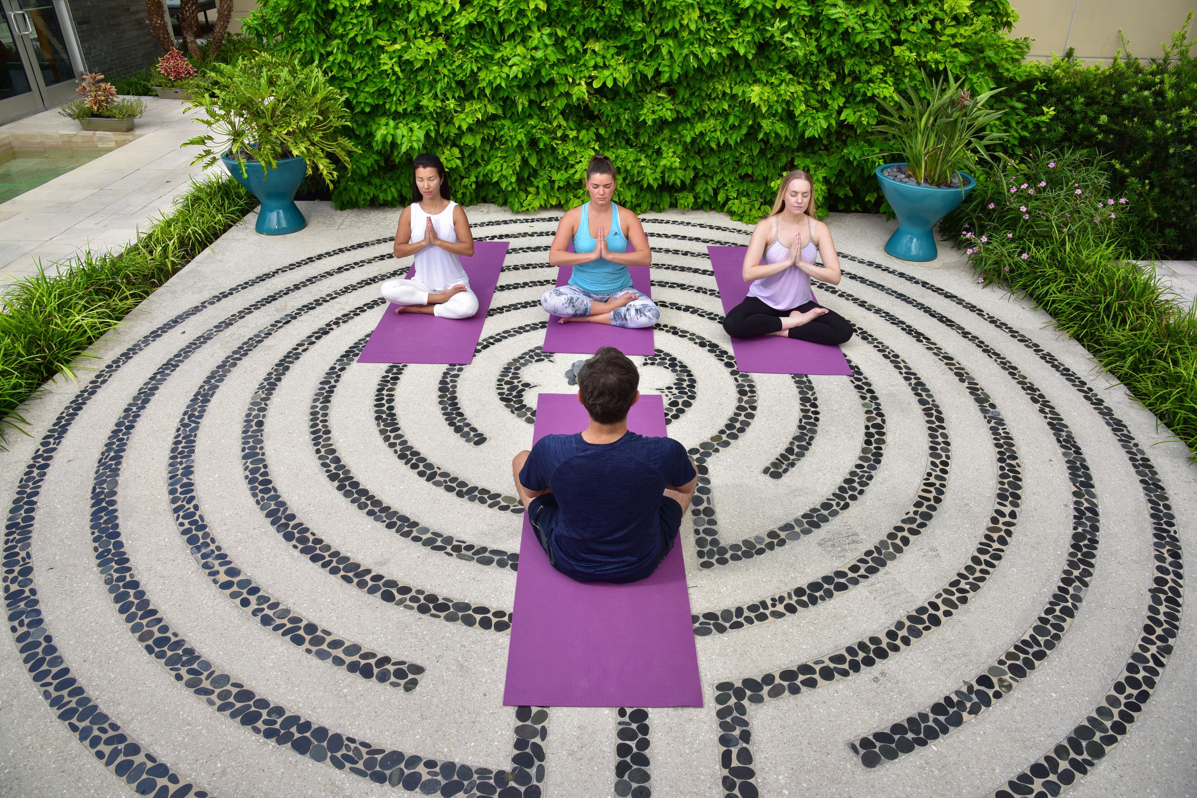 Drei Personen meditieren auf Yogamatten in einer kreisförmigen Anordnung auf einem gepflasterten Bereich mit Garten im Hintergrund.