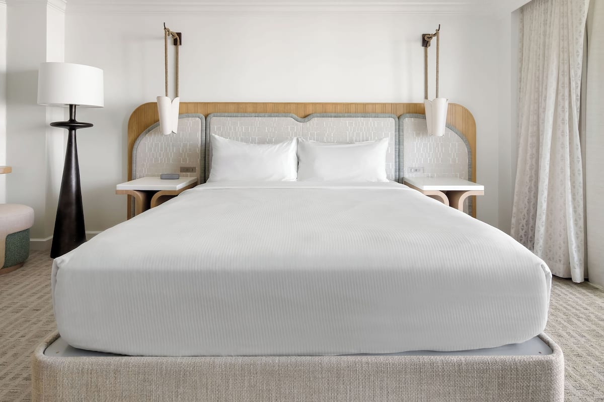 Una cama grande con cabecera de madera, dos mesitas de noche, una lámpara y una mesita con un florero.