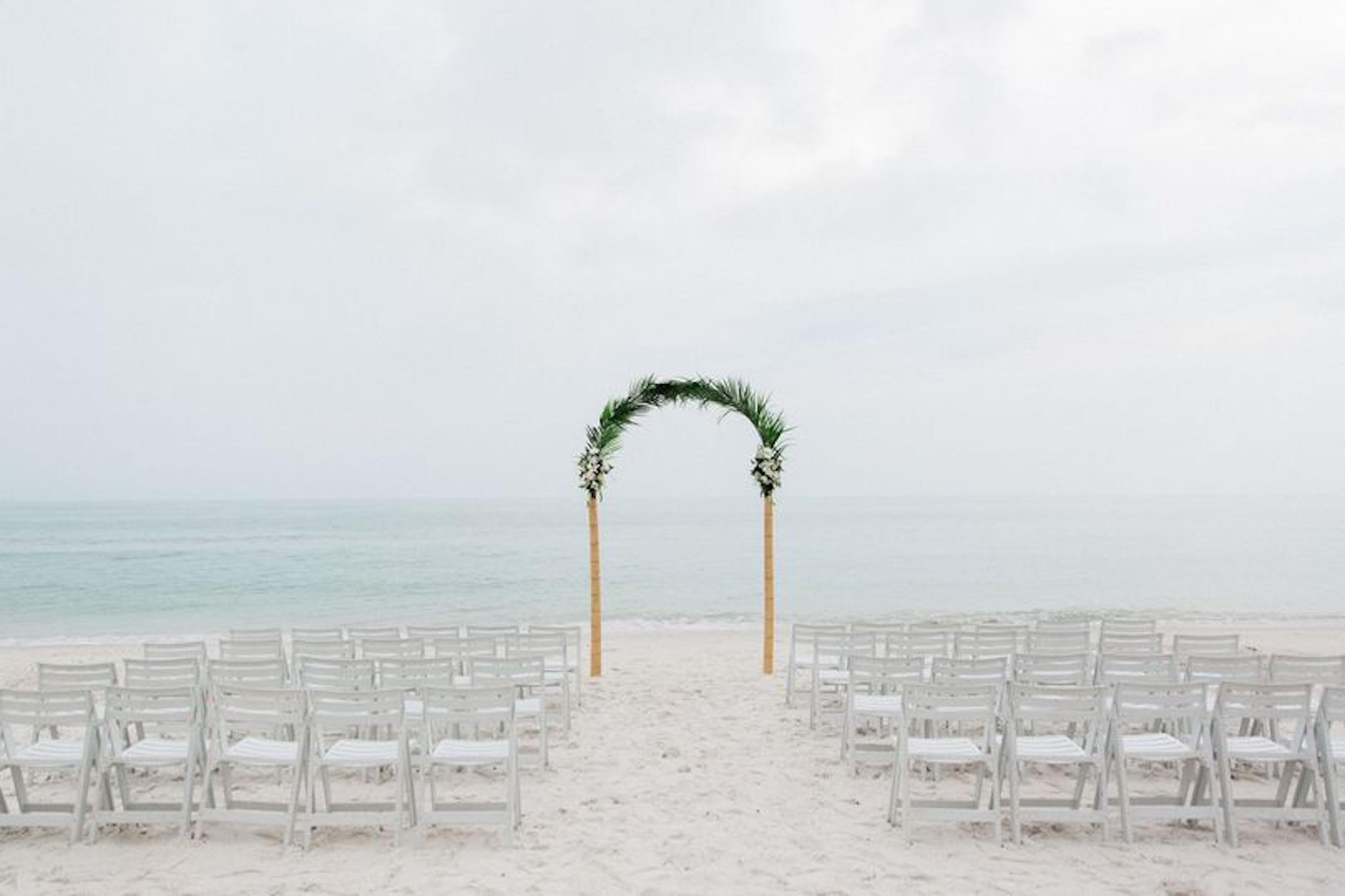 A beach wedding setup with white chairs and a floral arch.