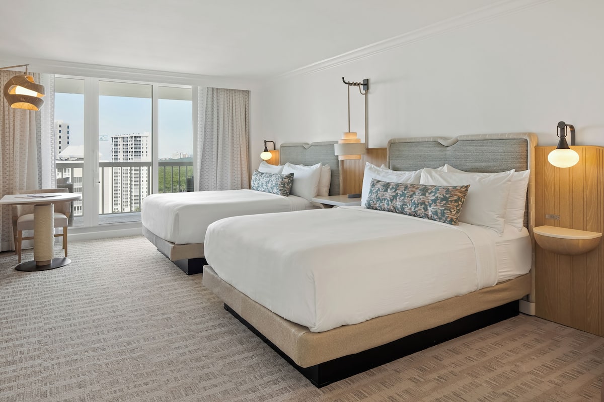 Habitación de hotel con dos camas, un escritorio, una silla, un ventanal con vistas a edificios y un balcón.
