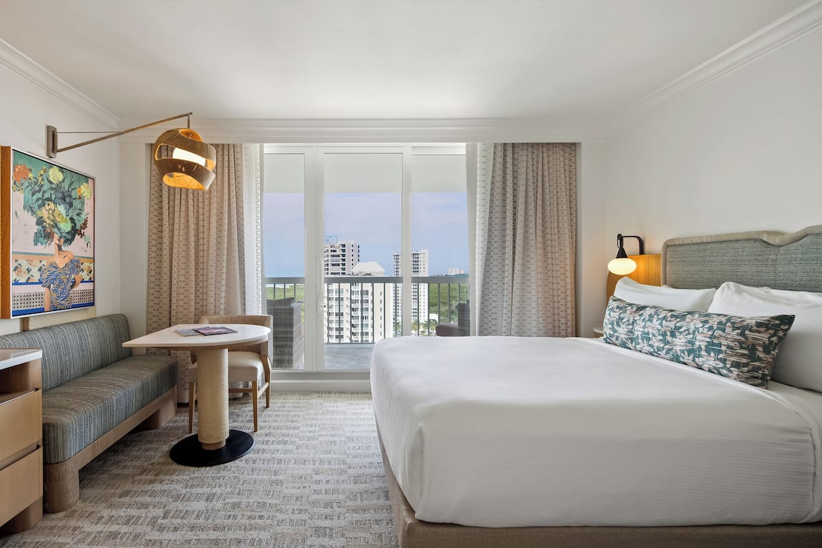 Habitación de hotel con cama, sofá, una mesita, una silla, una lámpara y un ventanal con vistas a la ciudad.