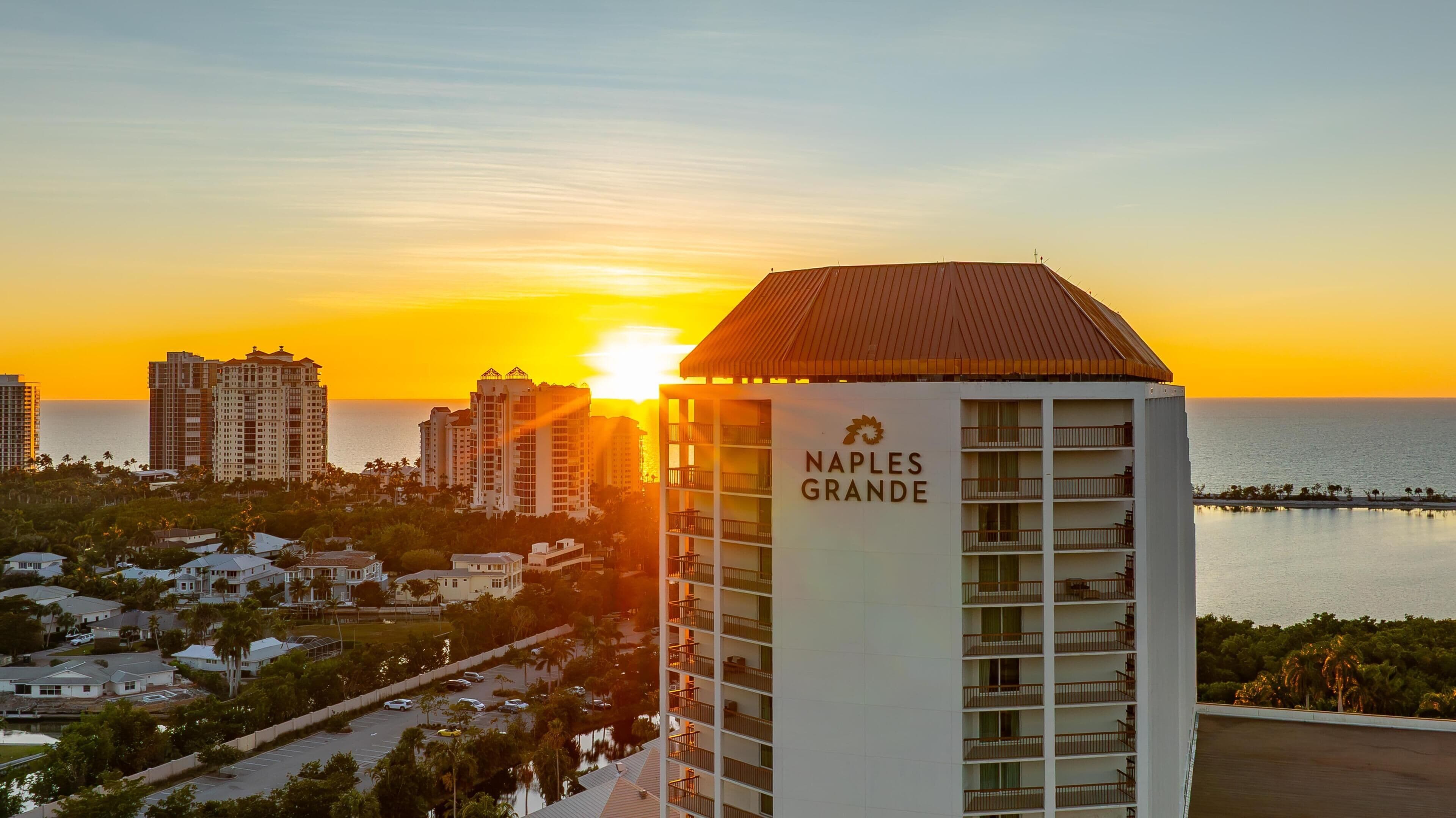 A high-rise hotel with a prominent sign 'NAPLES GRANDE' and a view of the sunset over the ocean.