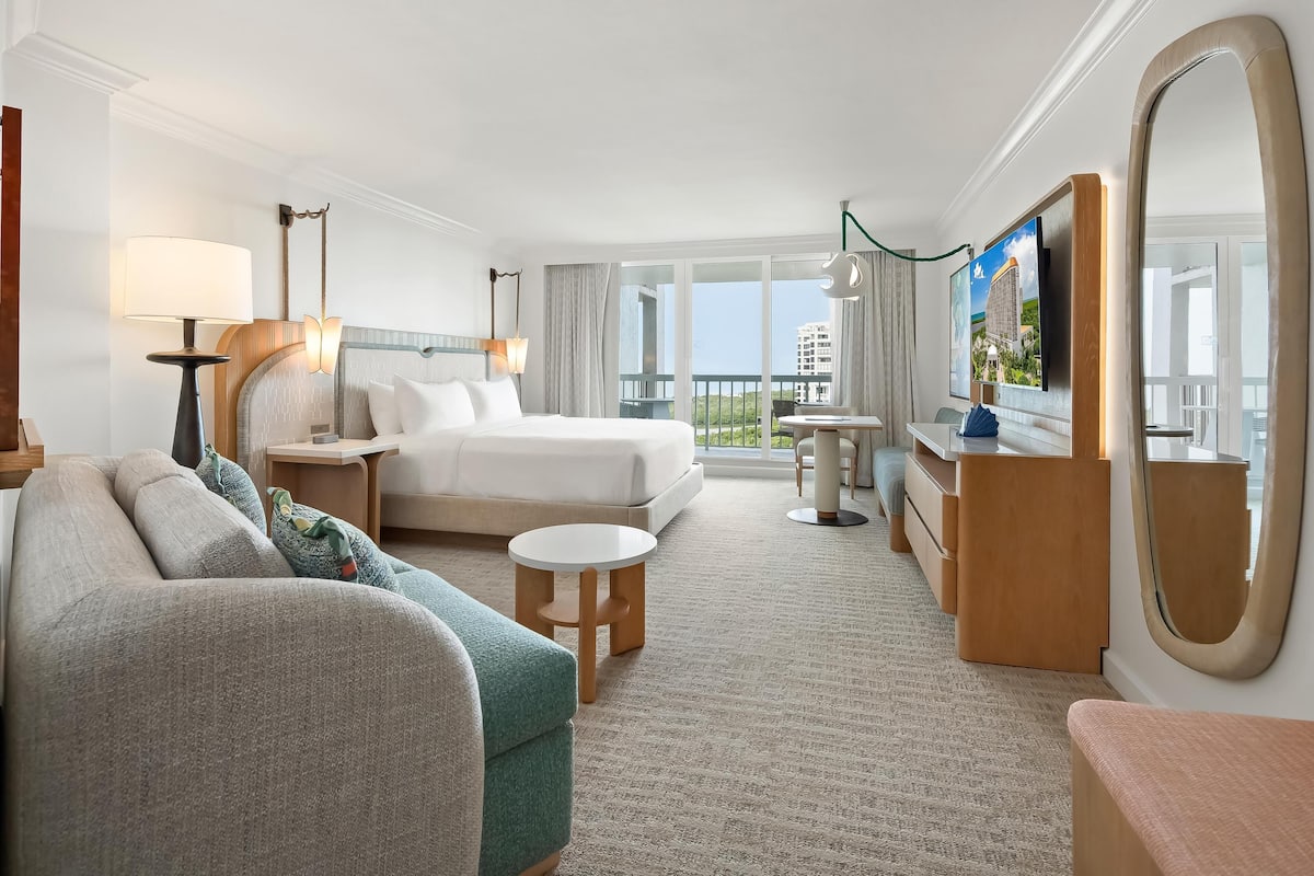 Habitación de hotel con cama, sofá, mesita, cómoda, televisor y vista al balcón.