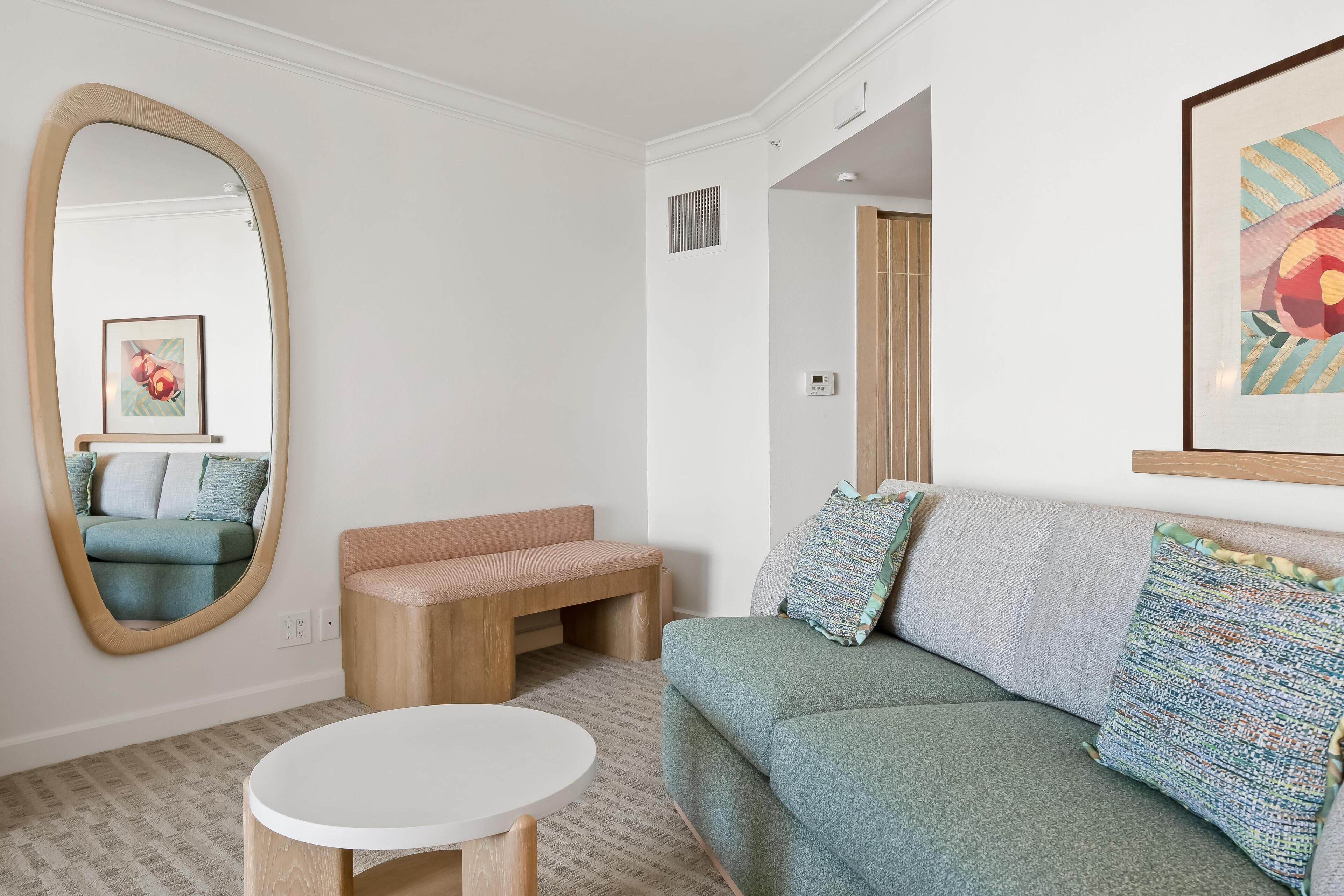 A modern living room with a large mirror, a round white table, a green sofa with patterned cushions, and a wooden bench.