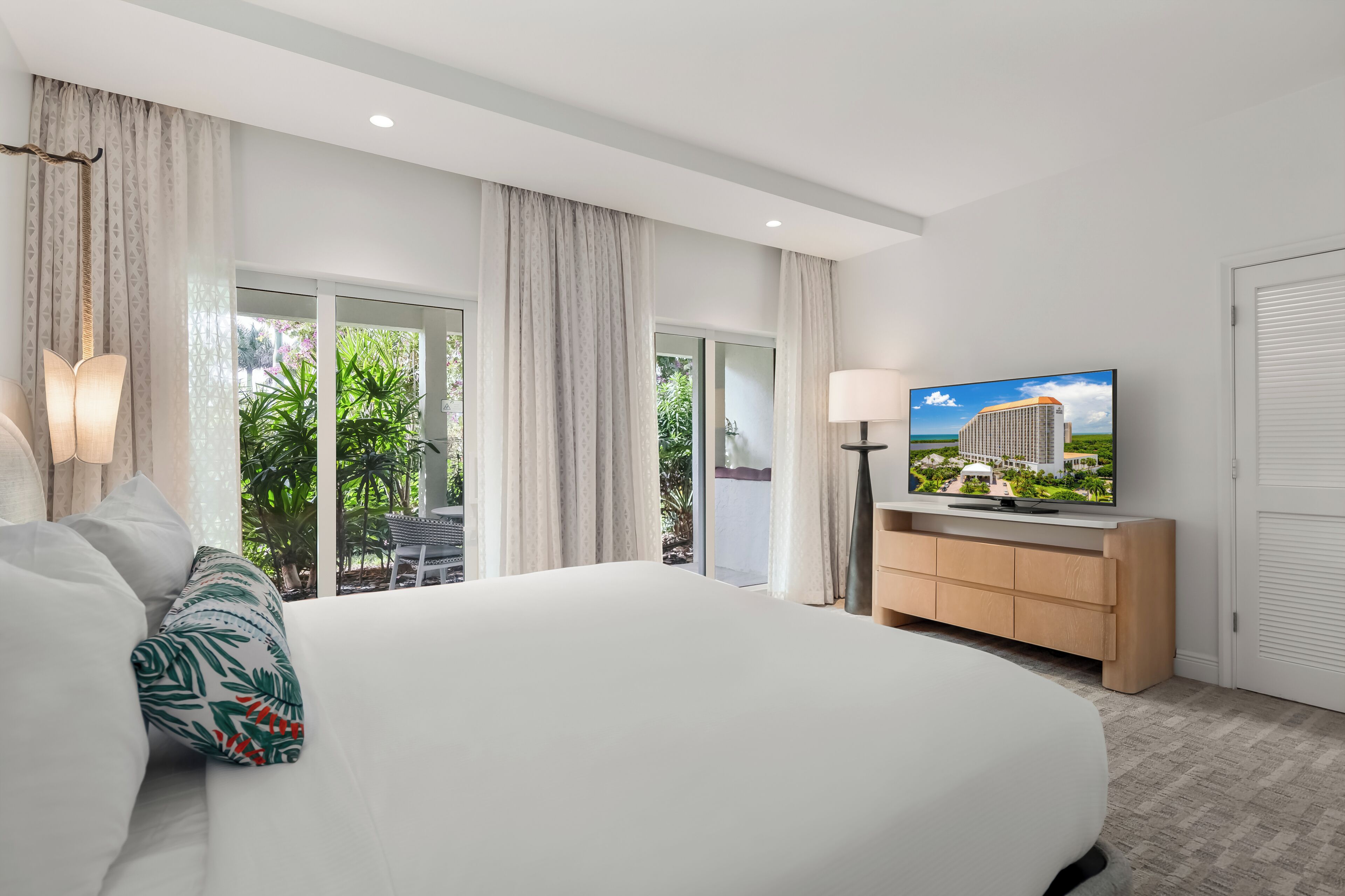 A hotel room with a large bed, a TV, a nightstand, and a view of greenery outside.