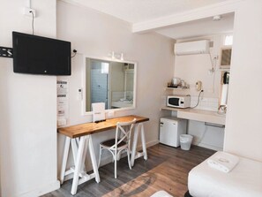 A compact room with a wooden desk, a white chair, a large mirror, a television mounted on the wall, a microwave, and a small refrigerator.