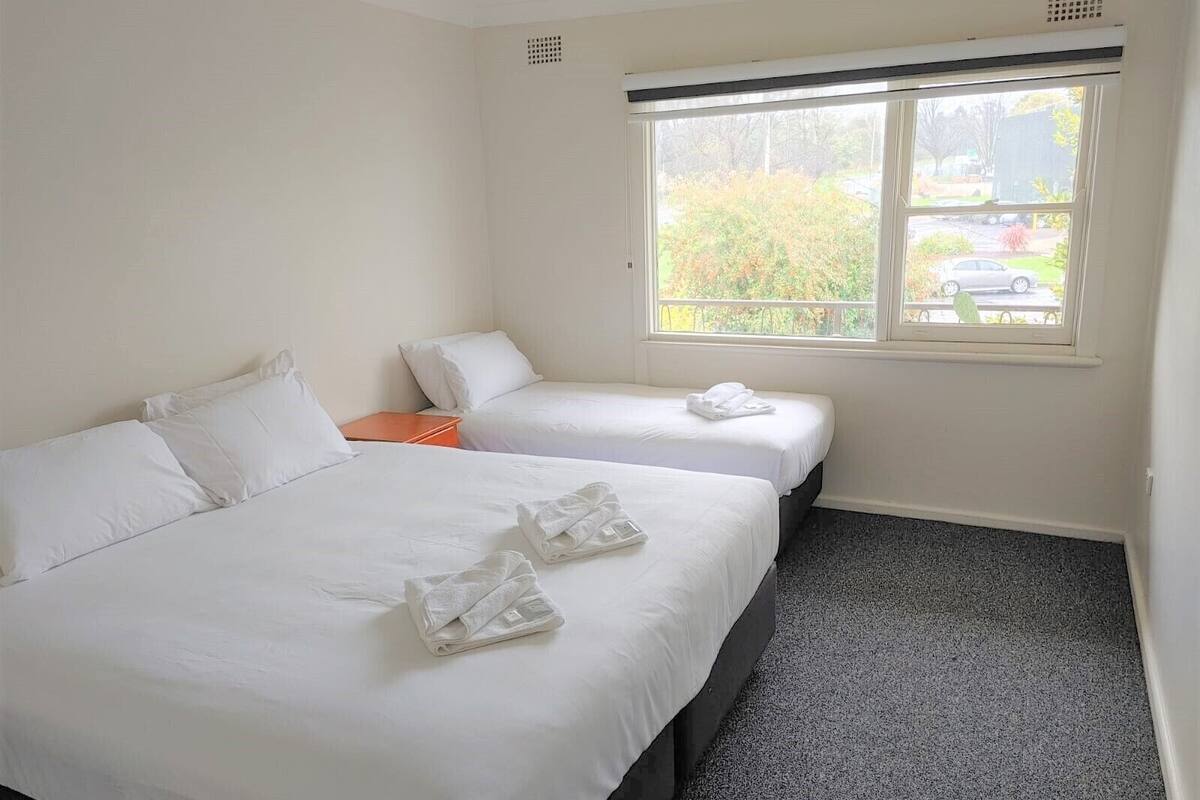 A hotel room with two beds, a window with a view of trees and a parking lot, and a carpeted floor.