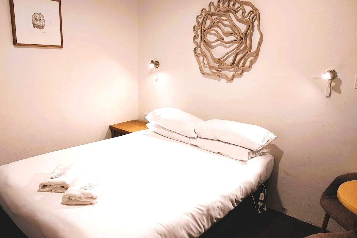 A neatly made bed with white linens and two pillows, a small wooden nightstand beside it, and a wall-mounted light fixture.