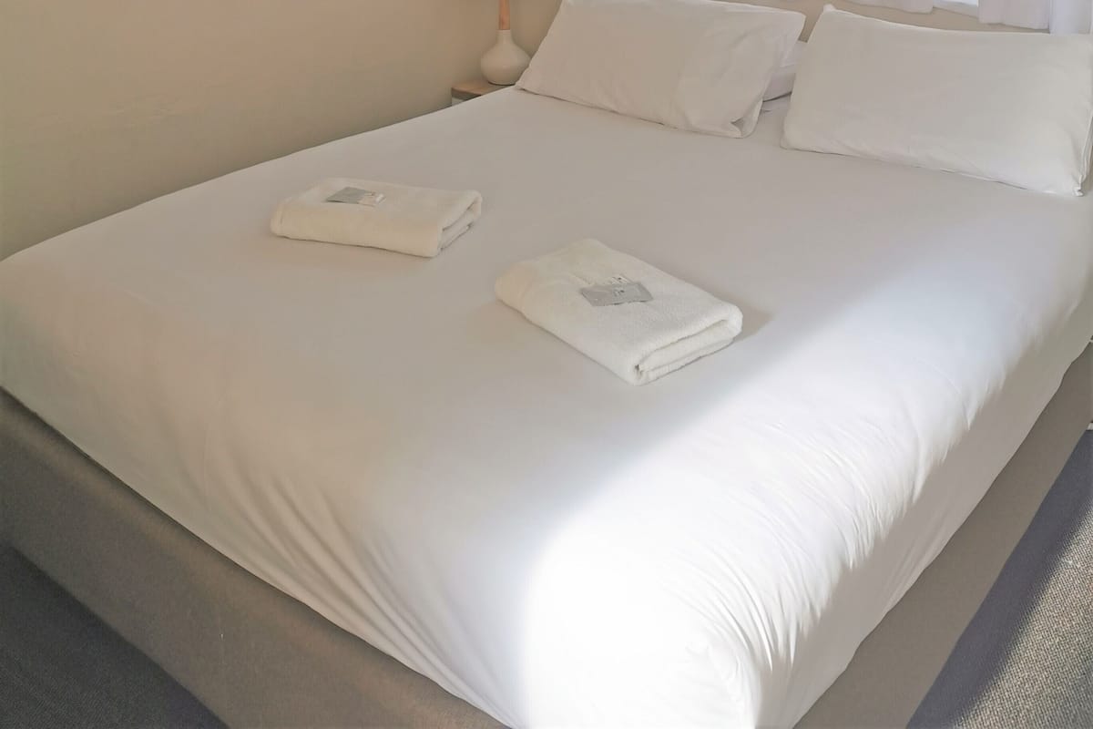 A neatly made bed with white linens and two folded towels, a bedside lamp, and a framed picture on the wall.