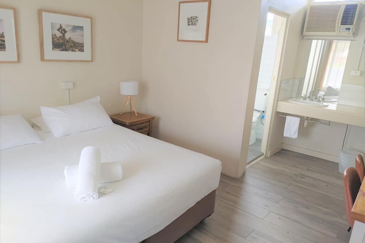 A hotel room with a large bed, a bedside table with a lamp, a bathroom with a sink and mirror, and framed pictures on the wall.